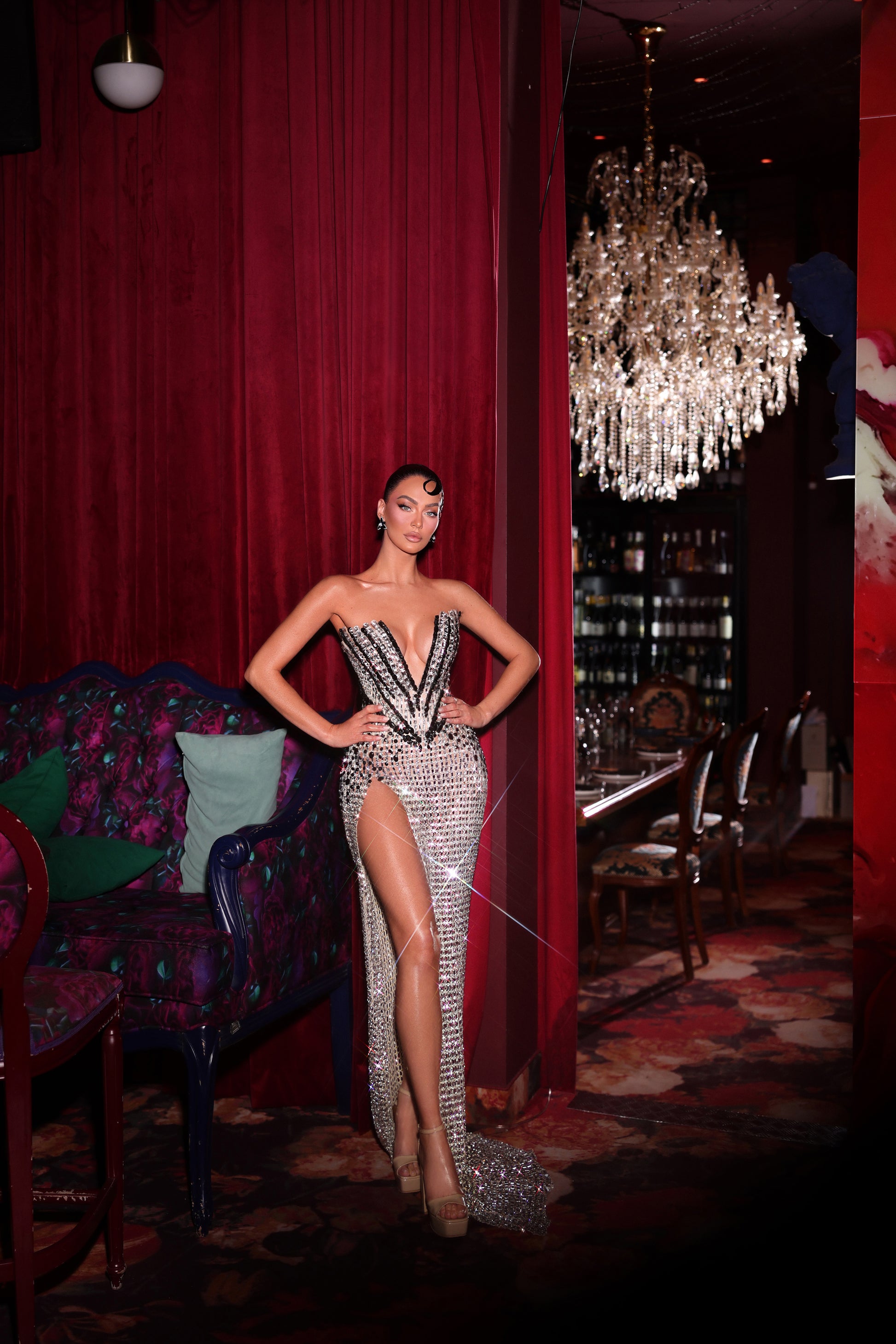 Woman in a sparkling dress standing in a glamorous room with red curtains and a chandelier.