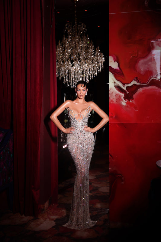Woman in a sparkling evening gown standing in front of a chandelier with red curtains.