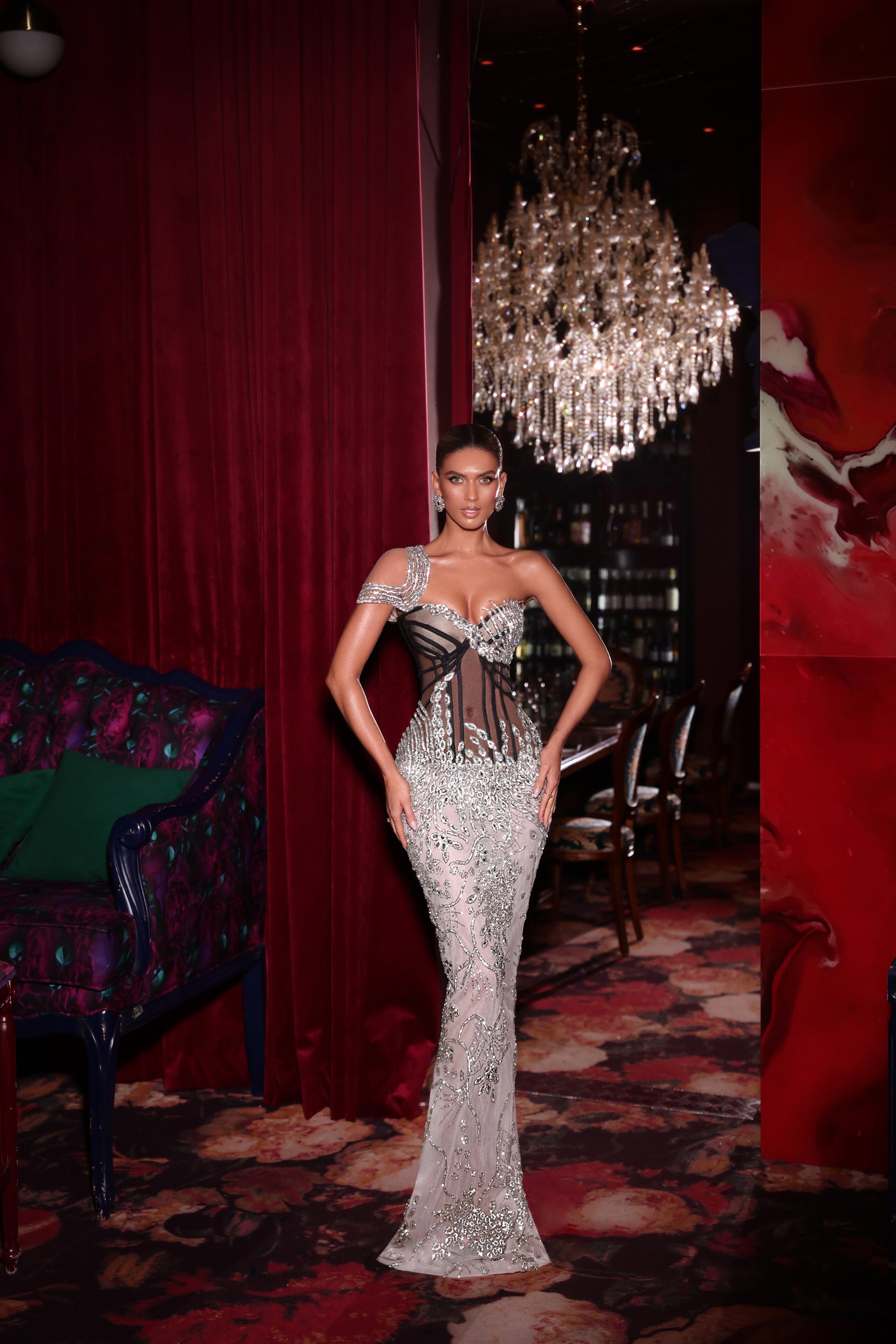 Woman in a silver evening gown standing in an opulent room with red curtains and a chandelier.