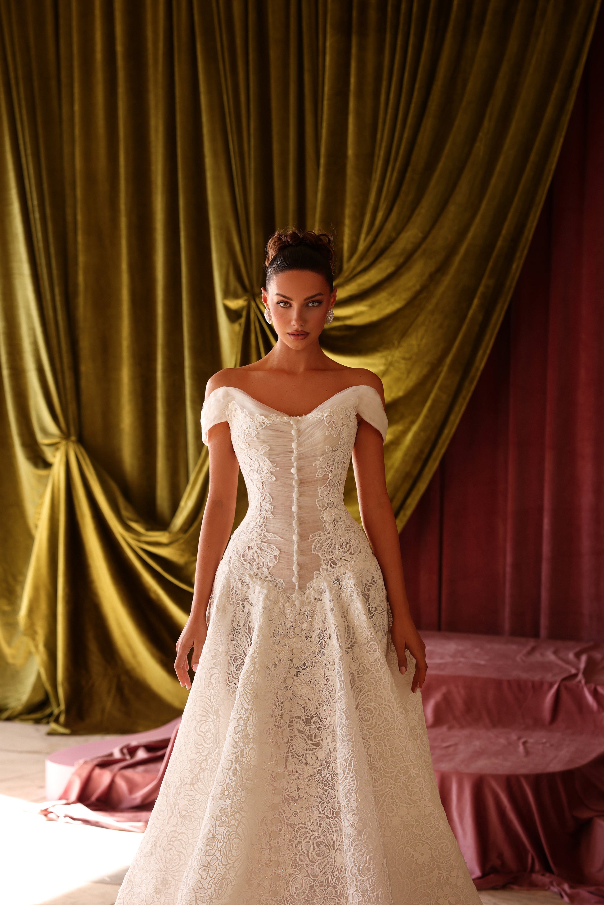 Model wearing a white lace wedding dress in front of draped curtains.