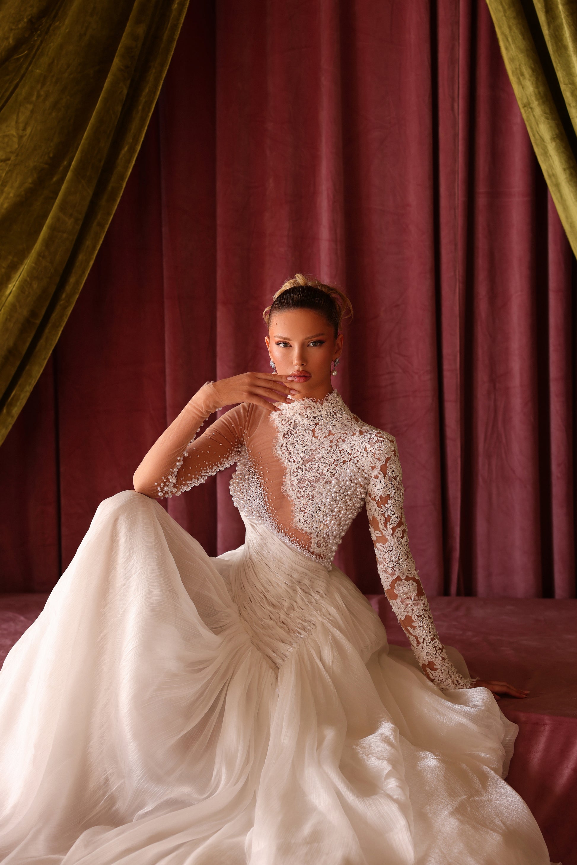 Woman in a white wedding dress with long sleeves and a high neckline, sitting against red curtains.