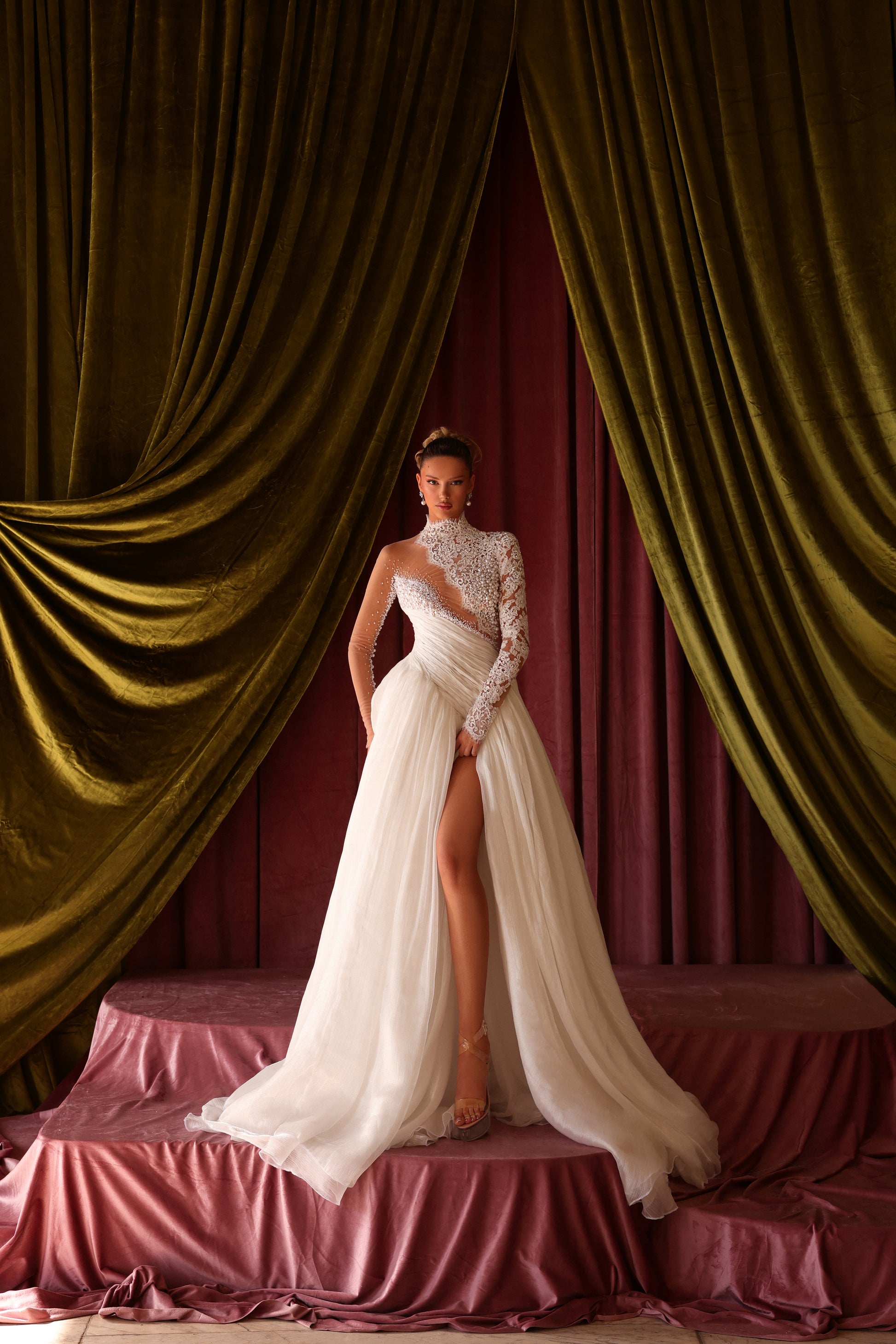 Woman in a white gown standing in front of draped green curtains.