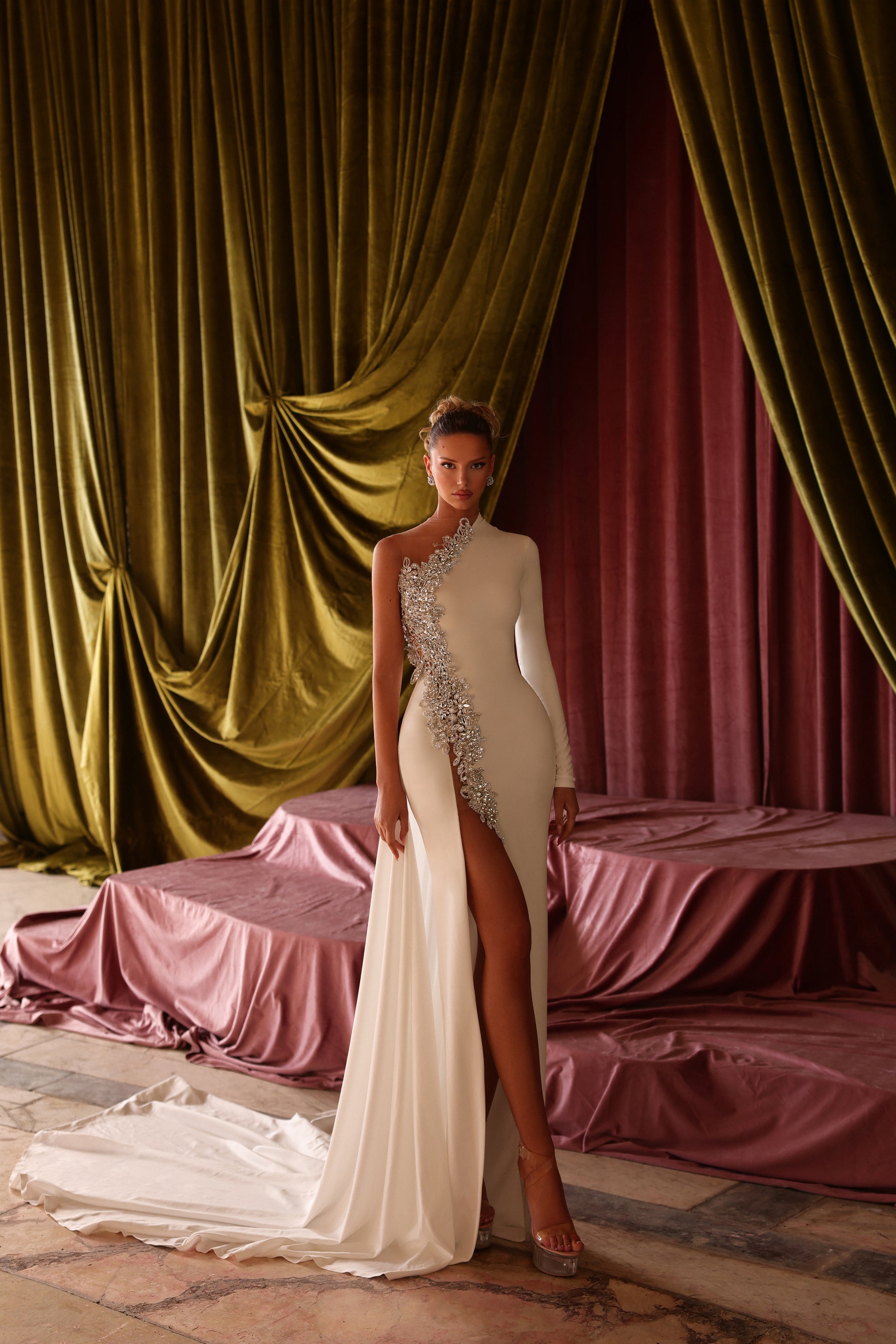 Woman in a white one-shoulder dress with a high slit standing in front of draped curtains.