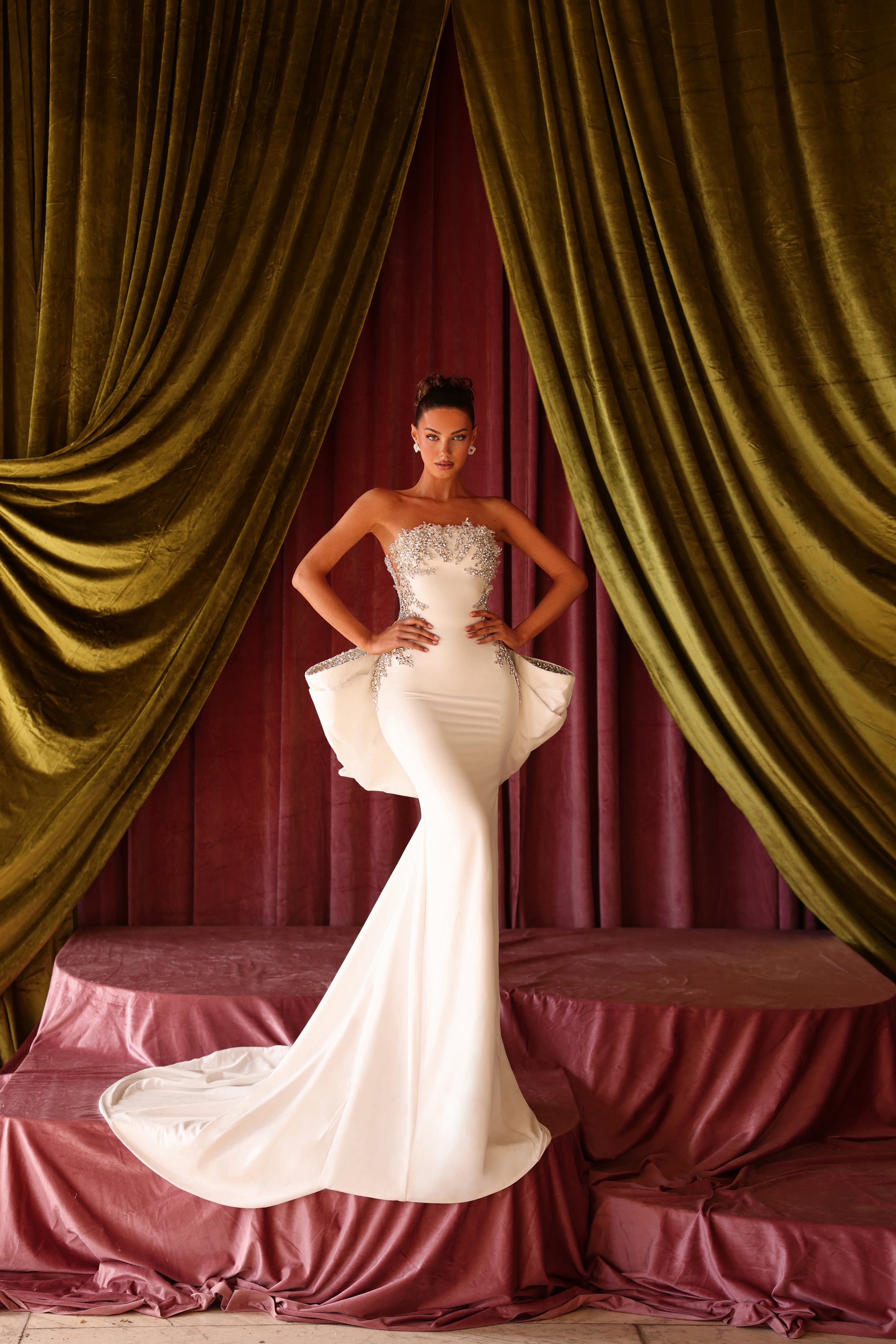 Woman in a white strapless gown standing in front of green velvet curtains.