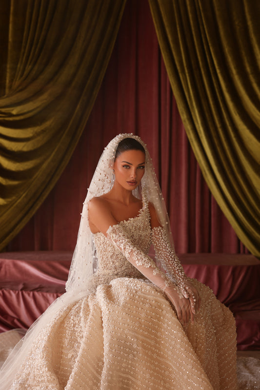 Woman in a wedding dress with a veil sitting in front of draped curtains.