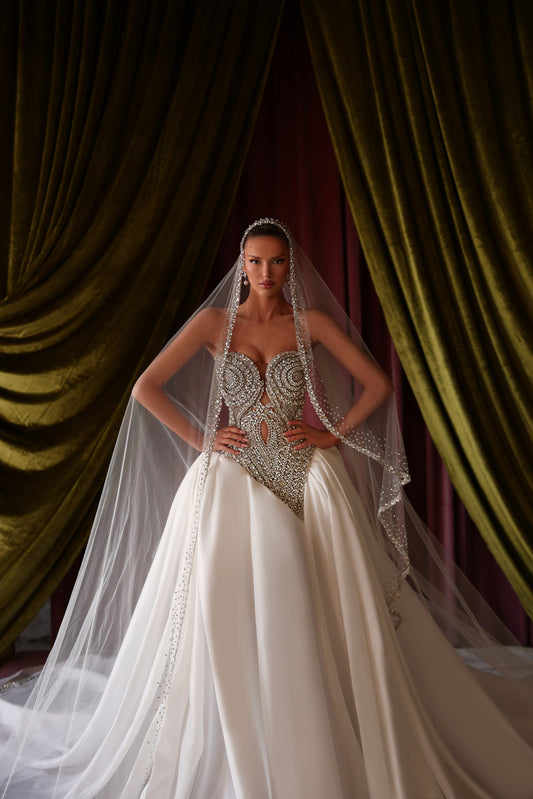 Woman in a white wedding dress with intricate detailing and a long veil against a draped curtain background.