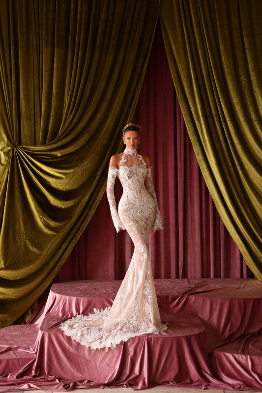 Woman in a white lace wedding dress standing in front of green and red draped curtains.