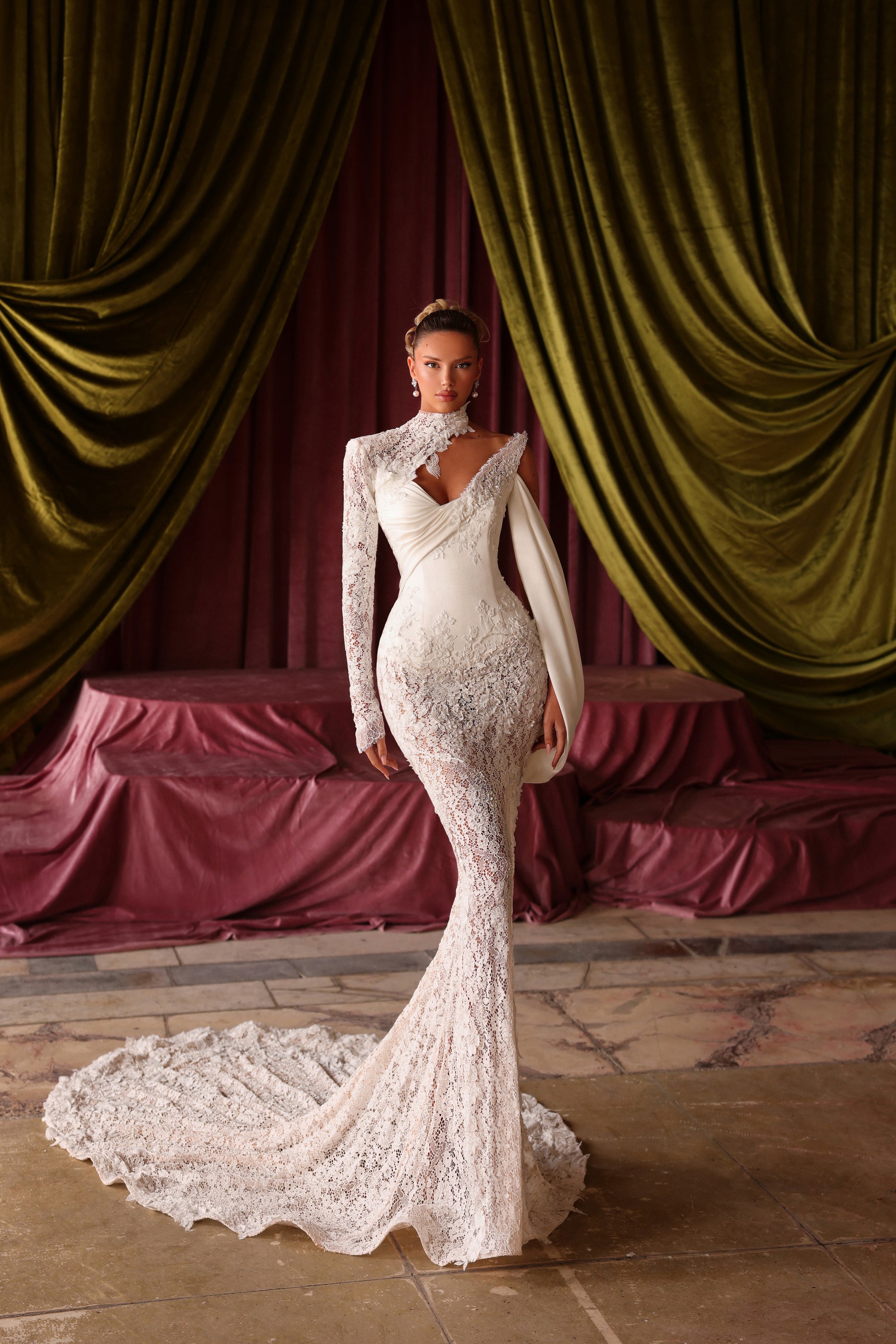 Woman in a white lace wedding dress standing in front of green velvet curtains.