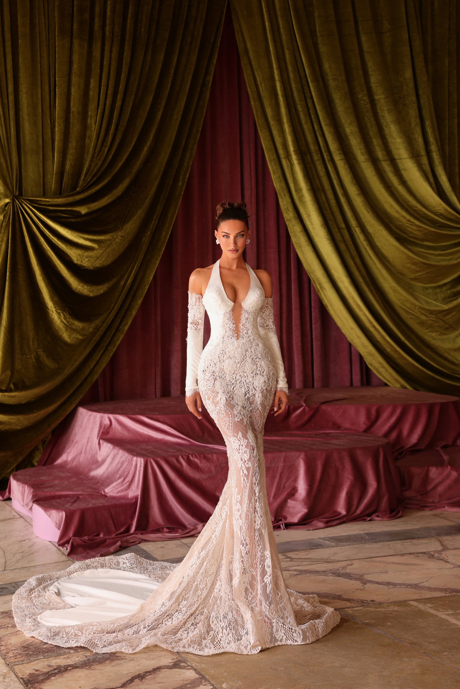 Woman in a white lace dress standing in front of green velvet curtains and a pink sofa.