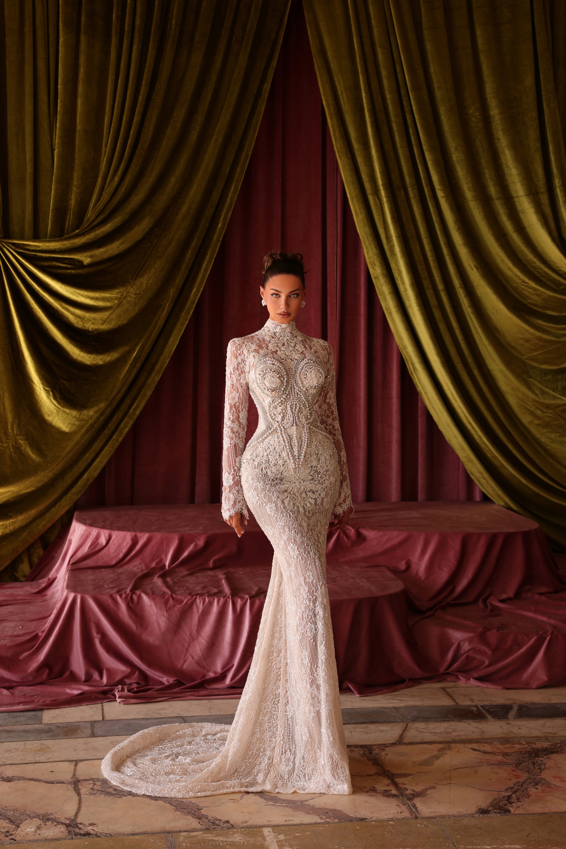 Woman in a white lace gown standing in front of gold curtains