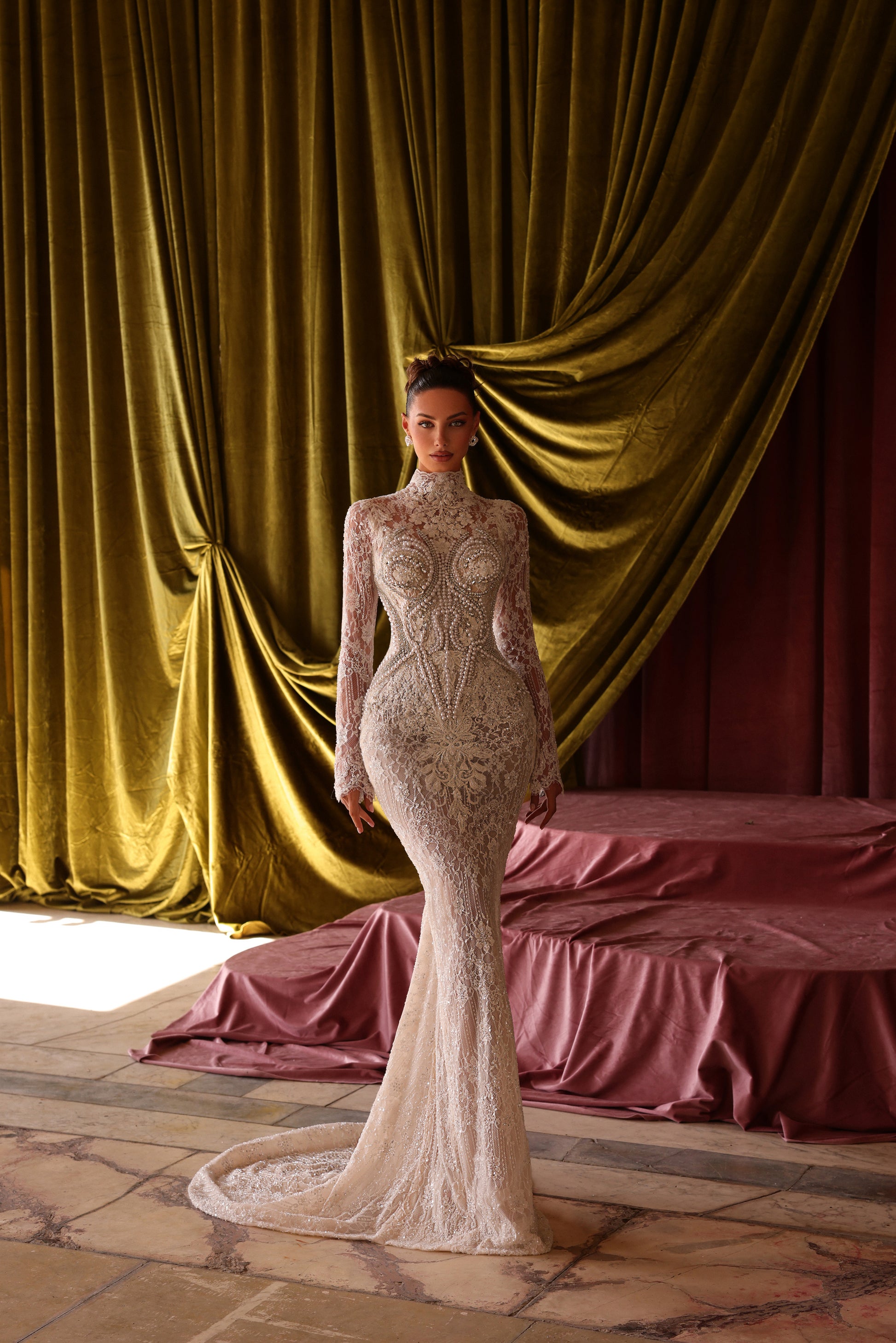 Woman in a detailed gown standing in front of gold curtains and a bed with pink bedding.