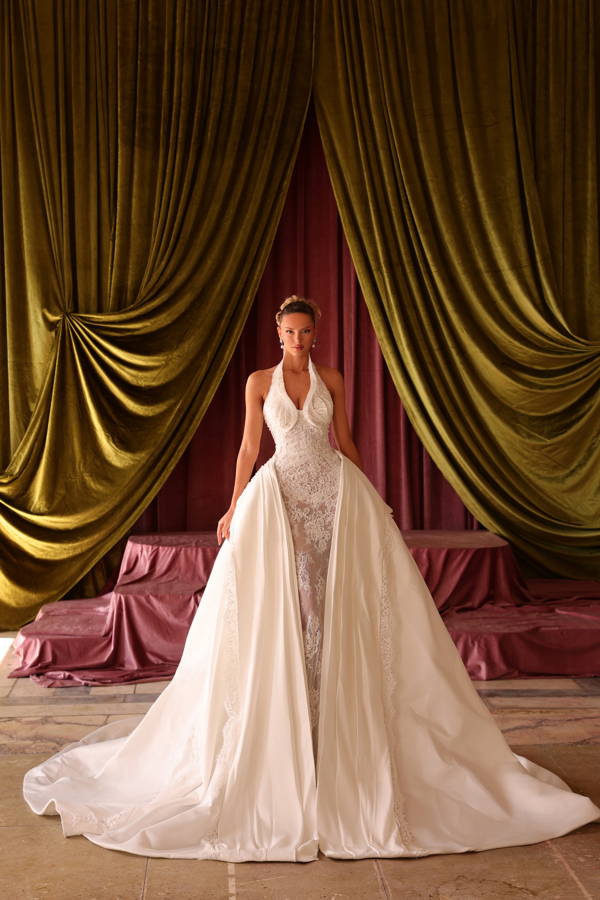 Woman in a white wedding dress standing in front of gold and red draped curtains.